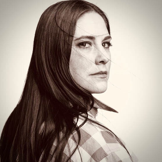 A sepia colored portrait of Jessica, a white woman with long, dark, straight hair.  A few flyaways blow in front of her face, which emotes a serious expression.  She sits in profile looking over her right shoulder and directly into the camera lens.  She wears a baggy, checkered man's shirt with a popped collar.
